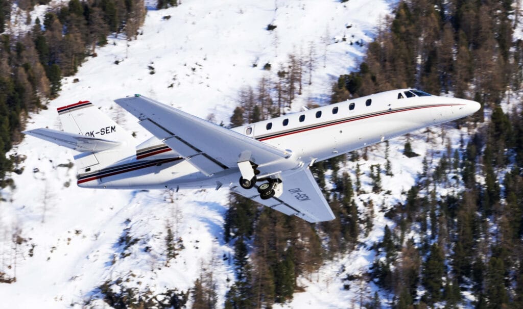 Ein Privatjet fliegt über schneebedeckte Berge, Bäume ragen aus dem Schnee. Auf dem Flugzeug steht "OK-SEM".