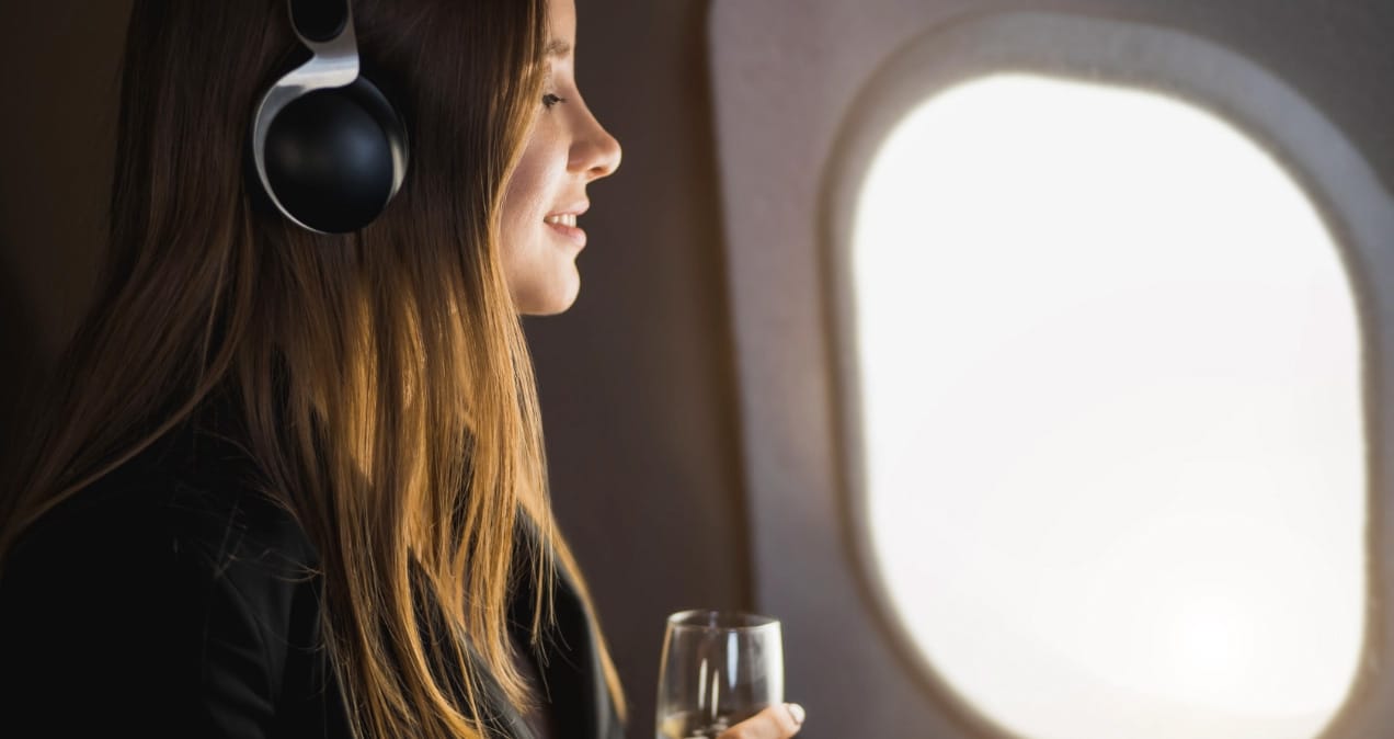Eine Frau mit Kopfhörern sitzt im Flugzeug, lächelt entspannt und hält ein Glas, während sie aus dem Fenster schaut.