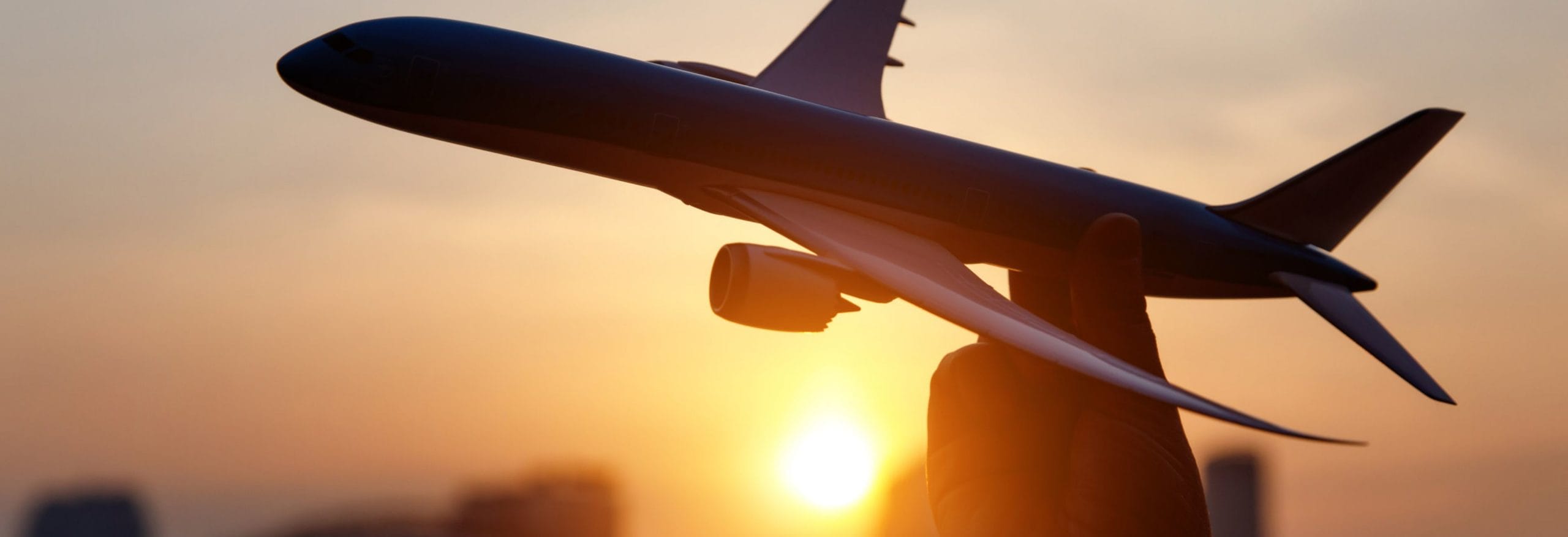 Eine Person hält ein Flugzeugmodell gegen einen sonnenuntergangsroten Himmel, im Hintergrund verschwommene Gebäude.