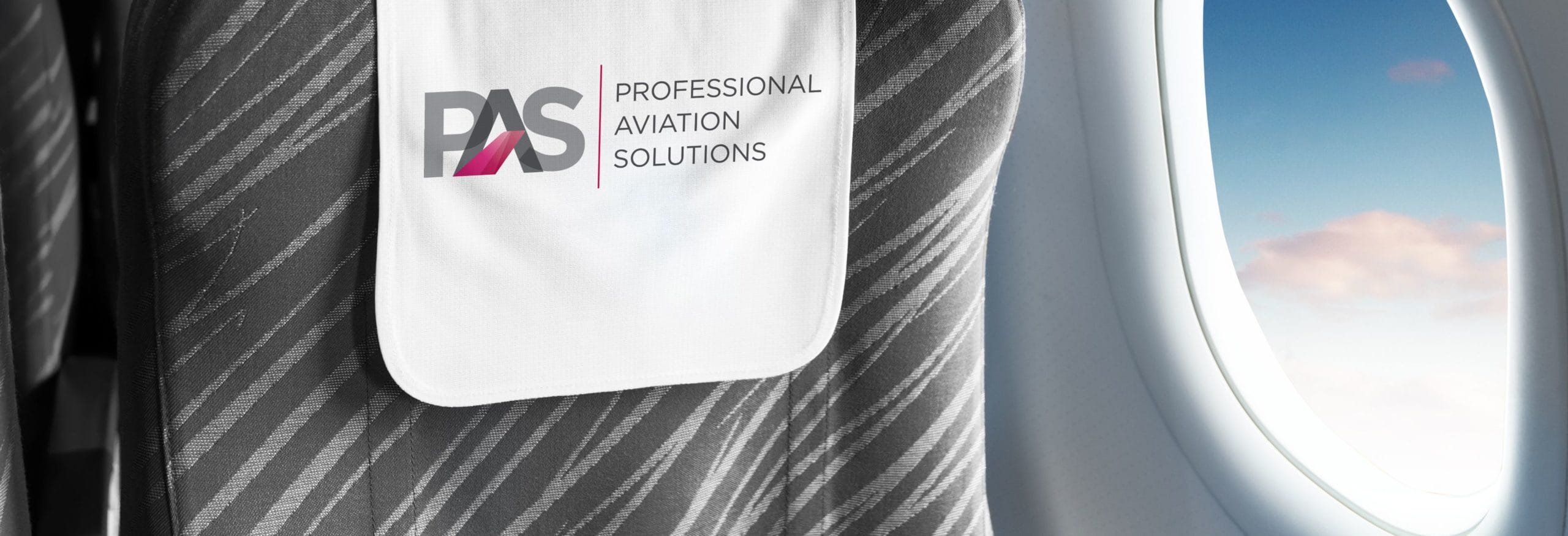 Ein grauer Flugzeugsitz mit weißem Kopfteil zeigt das Logo "PAS Professional Aviation Solutions" neben einem Fenster mit blauem Himmel sichtbar.