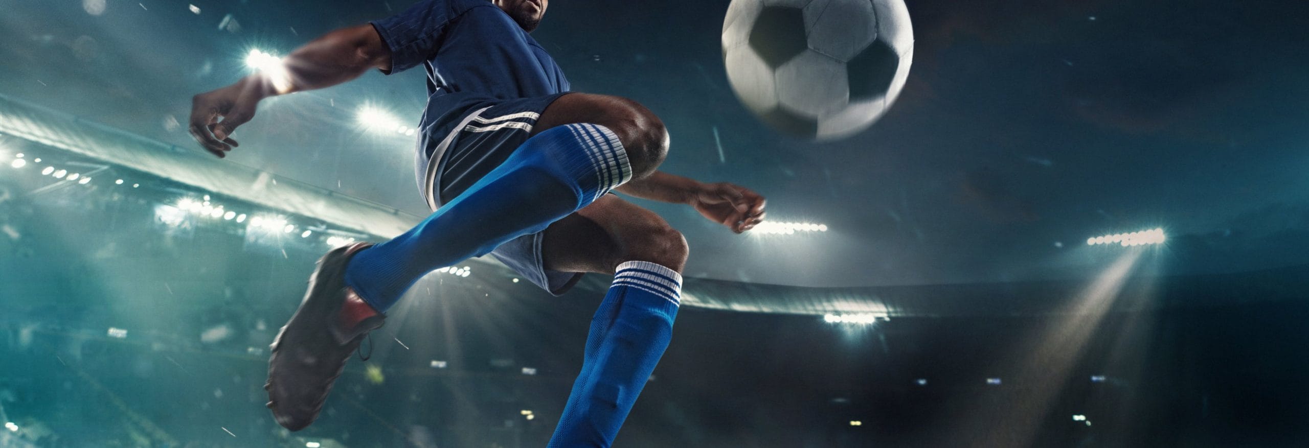 Ein Fußballspieler in blauer Uniform springt und tritt vor einem beleuchteten Stadion einen Fußball in der Luft.