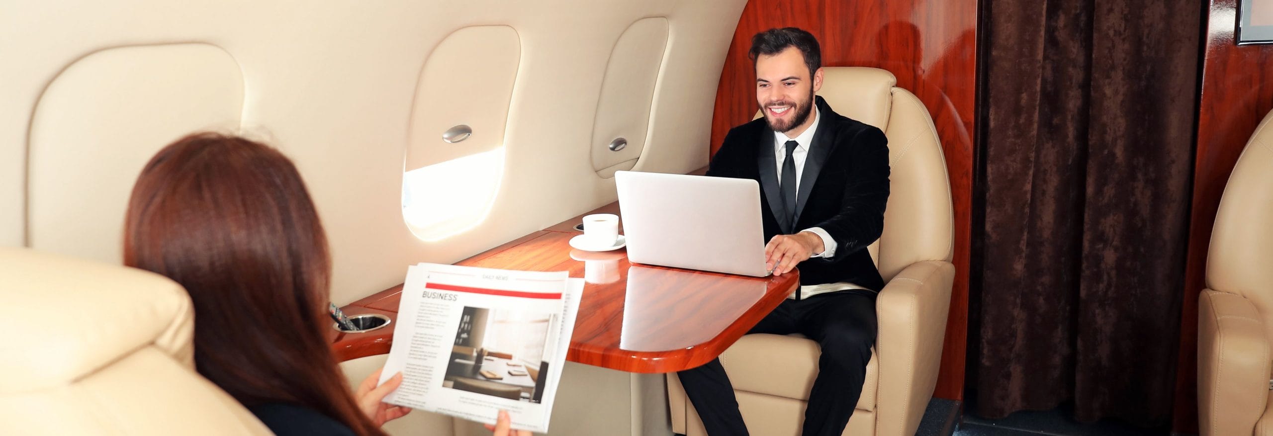 Eine Person arbeitet am Laptop an einem Holztisch in einem luxuriösen Privatjet. Im Vordergrund liest jemand eine Zeitung.