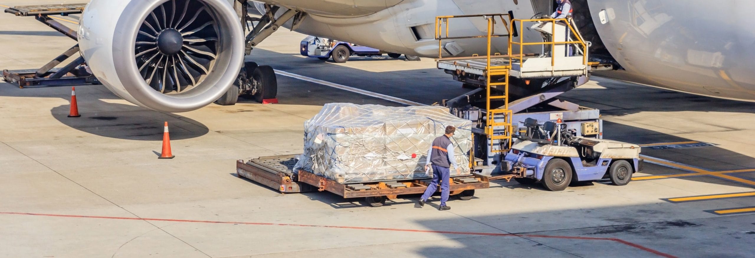 Ein beladenes Frachtfahrzeug transportiert verpackte Güter auf dem Rollfeld zu einem Flugzeug, während Arbeiter den Transport überwachen.