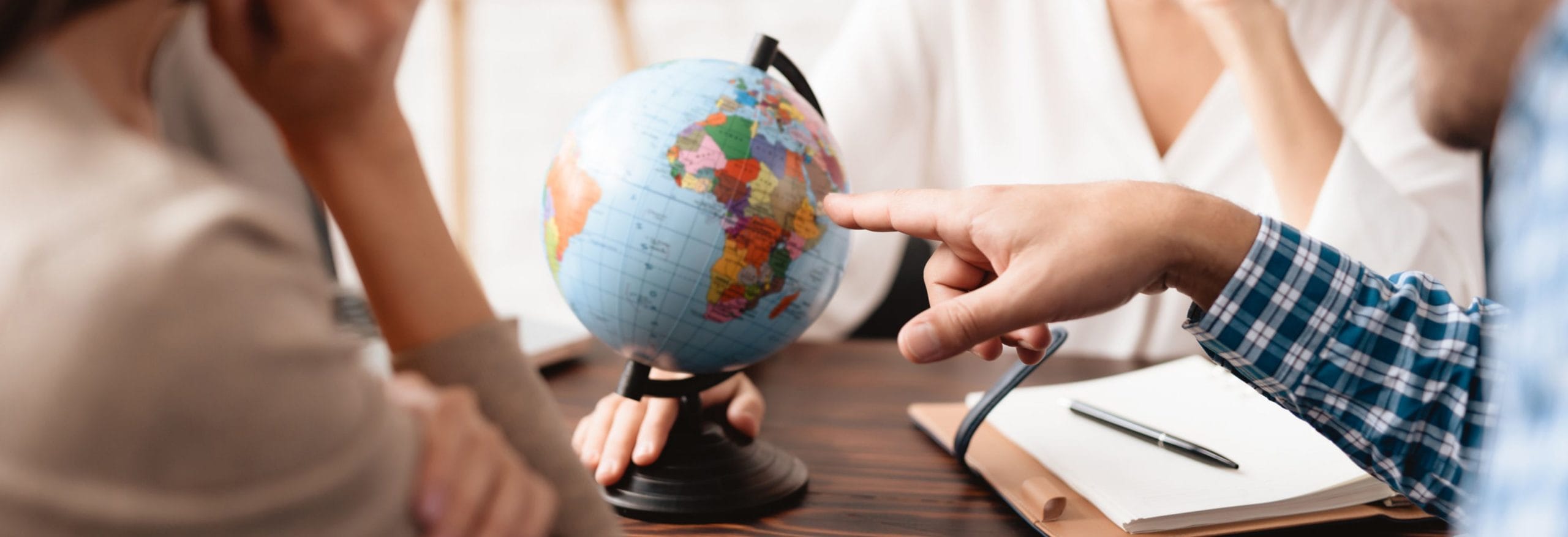 Ein Finger zeigt auf einen Globus auf einem Tisch. Um den Tisch sitzen mehrere Personen mit Schreibmaterialien.