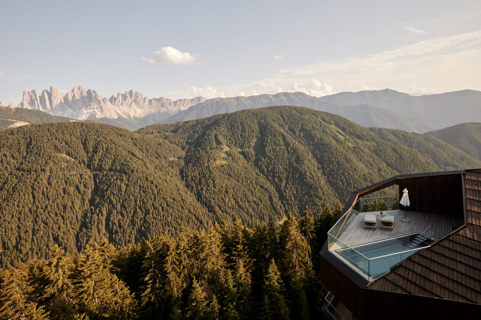 Liegestühle stehen unter Sonnenschirm auf Holzterrasse, überblicken Berge bei klarem Himmel.