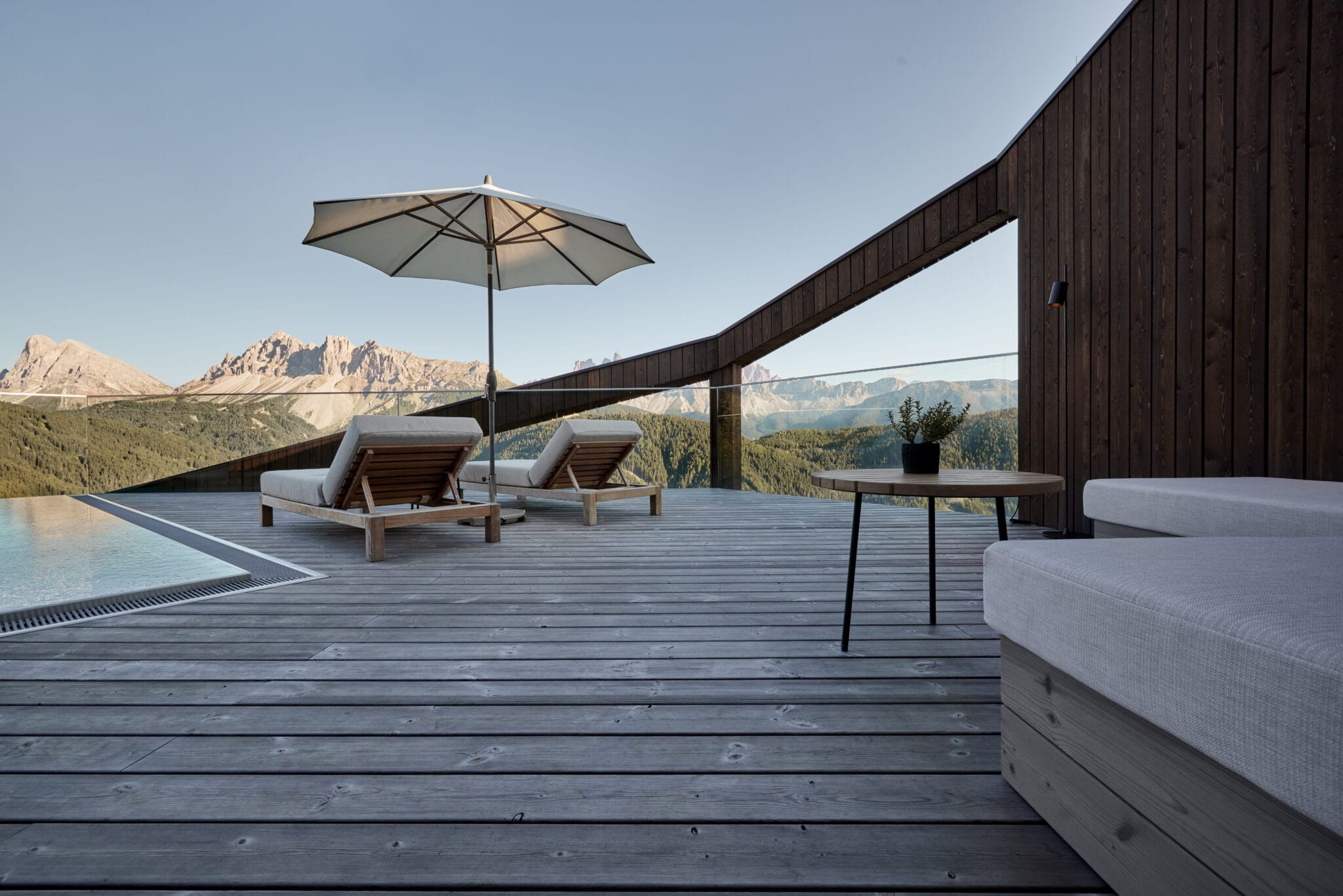Liegestühle stehen unter Sonnenschirm auf Holzterrasse, überblicken Berge bei klarem Himmel.