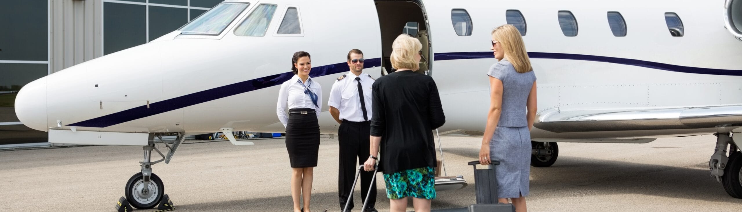 Eine Passagierin steigt mit einem Koffer in ein Privatflugzeug ein. Zwei Flugbegleiter und ein Pilot stehen zur Begrüßung bereit. Umgebung: Flughafen.