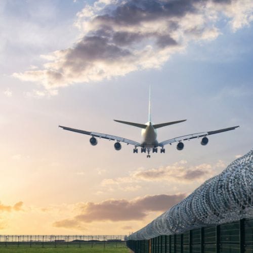 Ein Flugzeug landet am Himmel über einer grünen Wiese, begrenzt durch einen Maschendrahtzaun bei Sonnenuntergang.