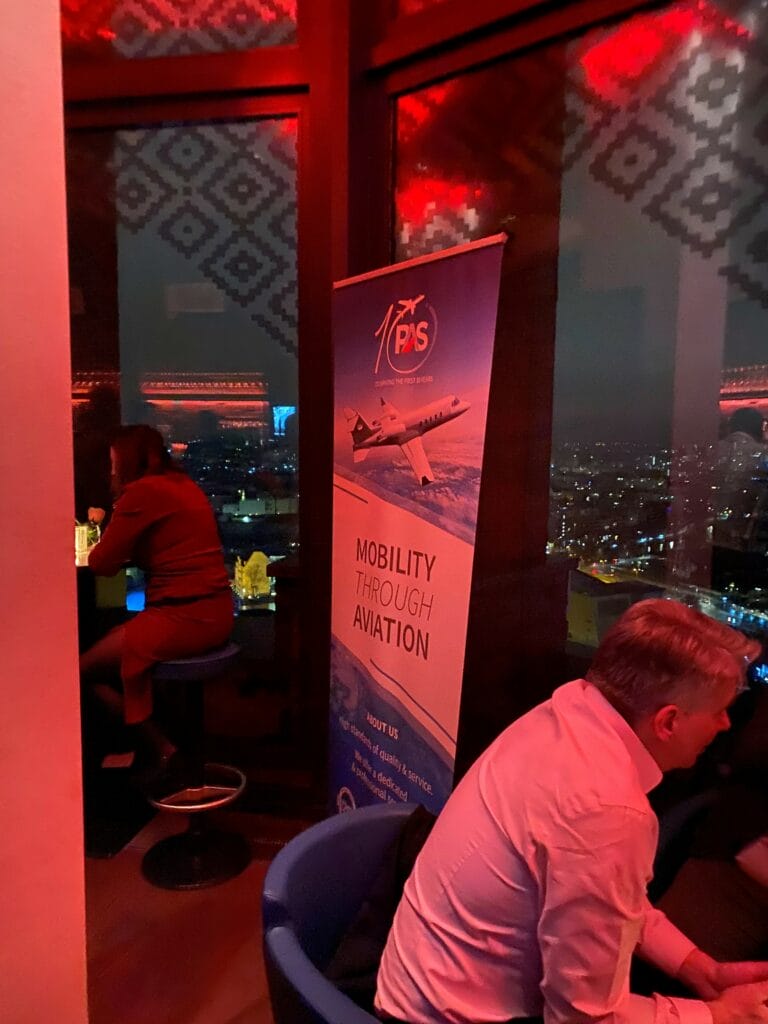 Ein Roll-Up-Banner zeigt ein Flugzeug mit der Aufschrift "Mobility Through Aviation". Menschen sitzen in einer rot beleuchteten Lounge mit Blick auf eine nächtliche Stadtlandschaft.
