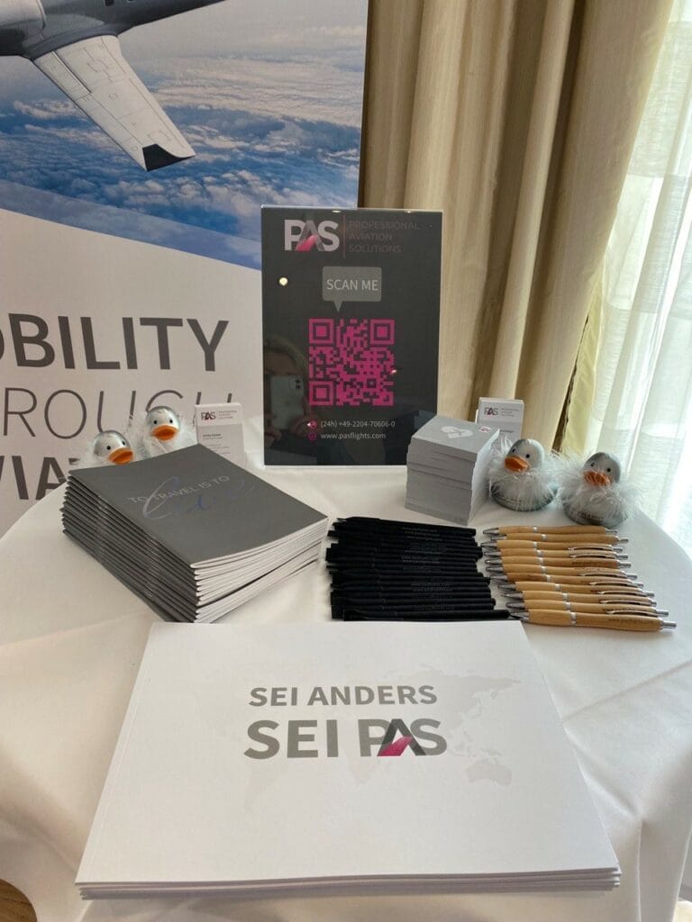 Büromaterialien liegen auf einem Tisch: Prospekte, Kugelschreiber, Plüschtiere. Ein Plakat zeigt ein Flugzeug am Himmel. Text: "SEI ANDERS SEI PAS" und QR-Code.