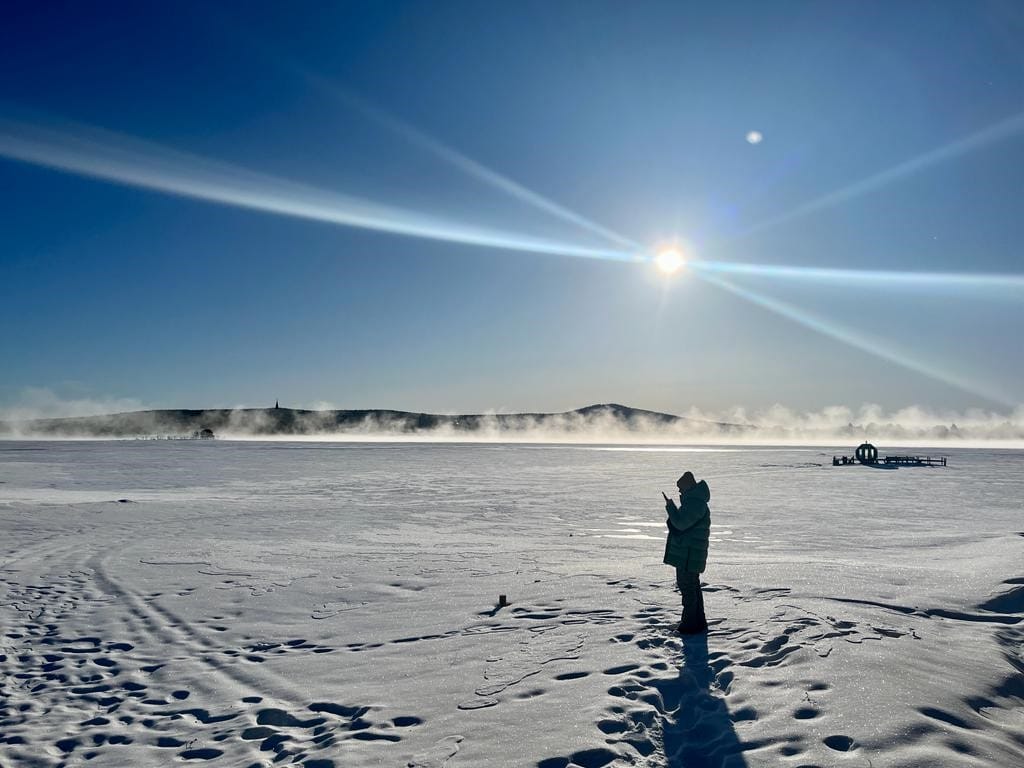 Person steht still in schneebedeckter Landschaft bei Sonnenschein und klarem Himmel, umgeben von Spuren auf dem Schnee und fernen Hügeln.