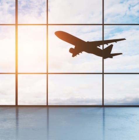 Ein Flugzeug fliegt hoch am Himmel, sichtbar durch eine verglaste Flughafen Fensterfront. Im Hintergrund sind Wolken und Sonnenlicht.