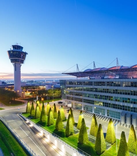 Ein Flughafen mit modernem Kontrollturm steht beleuchtet in der Abenddämmerung. Im Vordergrund sind geometrisch angeordnete Bäume zu sehen.