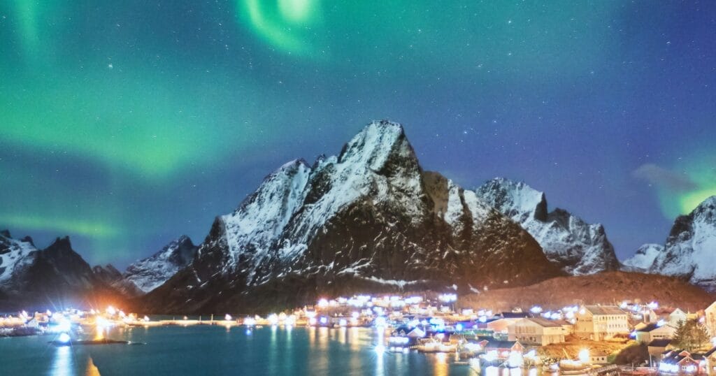 Nordlichter tanzen über schneebedeckten Bergen, erhellen ein malerisches Dorf am Wasser; spektakuläres Naturschauspiel vor winterlicher Kulisse.