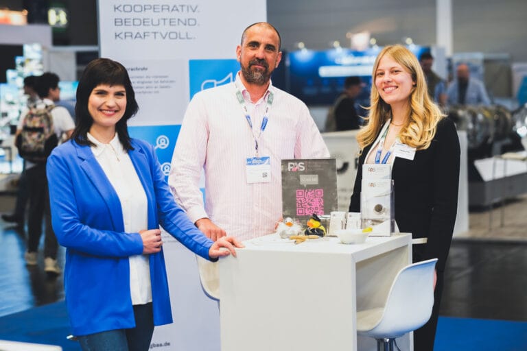 Drei Personen lächeln an einem Messestand. Im Hintergrund ist ein Roll-up mit Text: „KOOPERATIV. BEDEUTEND. KRAFTVOLL.“. Der Stand enthält Visitenkarten und Dekoration.