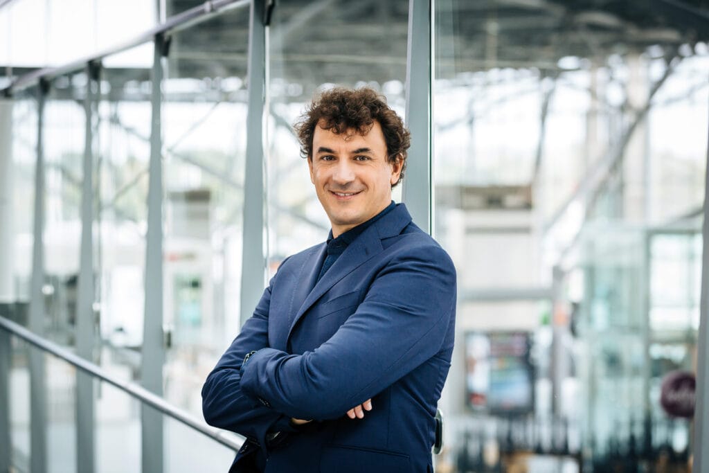 Alessio Altamura, Managing Director for PAS Italy, smiling in a modern office environment. He is wearing a dark blue suit and stands with his arms crossed in front of a glass facade.