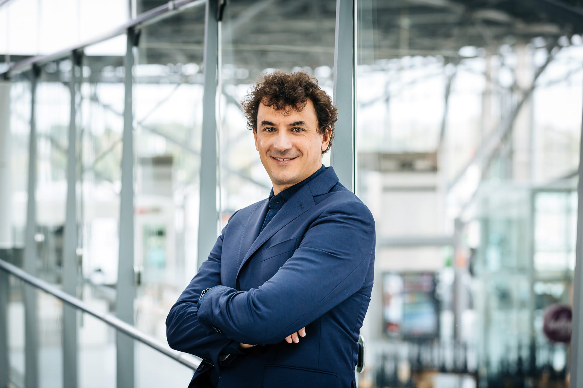 Alessio Altamura, Managing Director for PAS Italy, smiling in a modern office environment. He is wearing a dark blue suit and stands with his arms crossed in front of a glass facade.